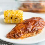 A whole baked chicken breast covered with bbq sauce on a white plate with more chicken in background