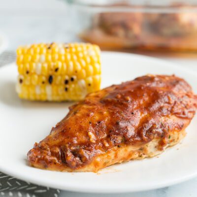 A whole baked chicken breast covered with bbq sauce on a white plate with more chicken in background