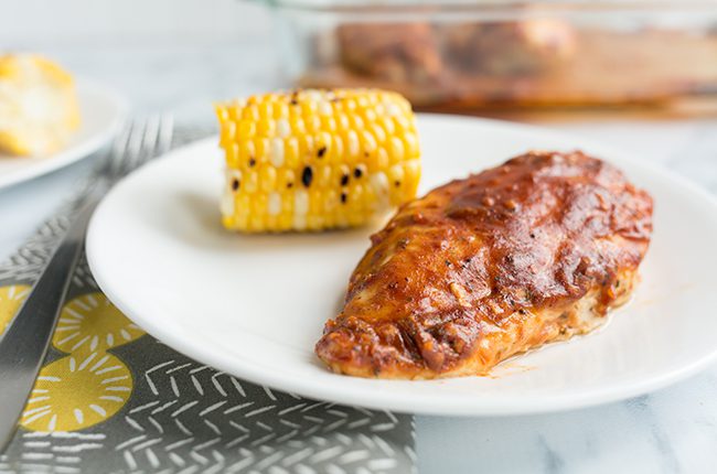White plate of bbq baked chicken with grilled corn sitting on a gray and yellow cloth napkin