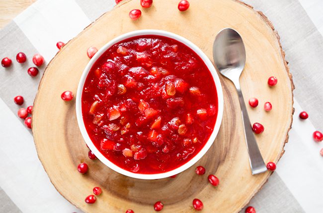 A white bowl filled with prepared cranberry apple chutney relish sitting on a slice of wood surrounded by fresh cranberries