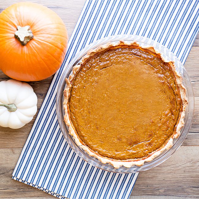 A whole baked healthy pumpkin pie sitting on a white and blue cloth next to mini pumpkins