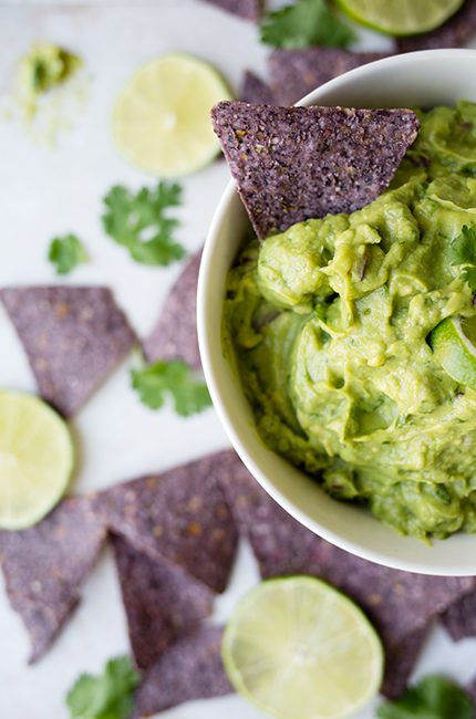 Picture of a white bowl filled with guacamole on a table with blue corn tortilla chips around the bowl