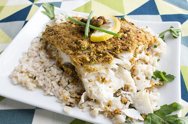 Pistachio crusted baked halibut sitting on a white plate with lemon on top