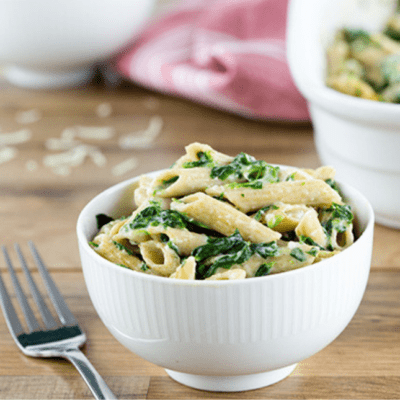 A large white bowl filled with spinach pasta and a fork on the table by the bowl