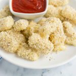 An up close picture of a white plate filled with baked mozzarella bites served with cup of marinara sauce