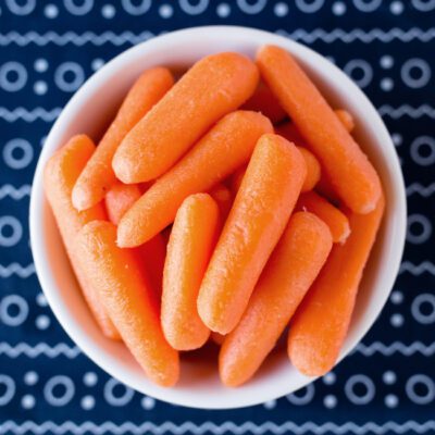 Square overhead picture of a white bowl of candied carrots sitting on a blue and white cloth