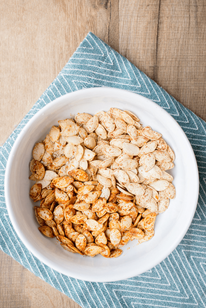 Overhead picture of a wooden table with a blue and white cloth that is topped by a bowl of pumpkin seeds